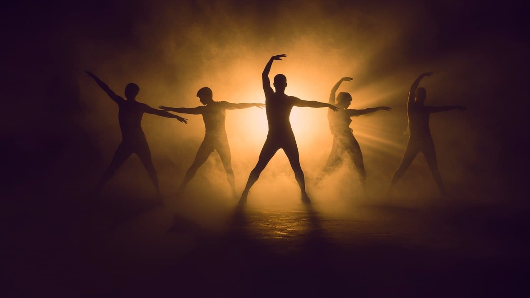 Five dancers in silhouette across a stage, mid-choreography, against strong backlight and fog.
